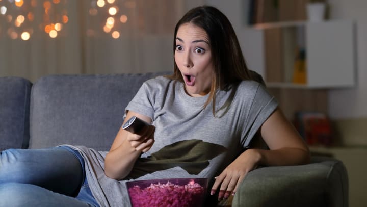 A woman on a couch with a TV remote in her hand, eating popcorn, looking surprised. 