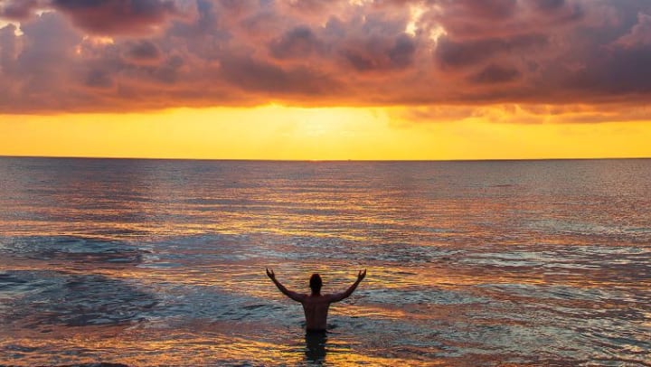 Man, waist-deep in the ocean with his arms outstretched, looking at the sunset in the distance