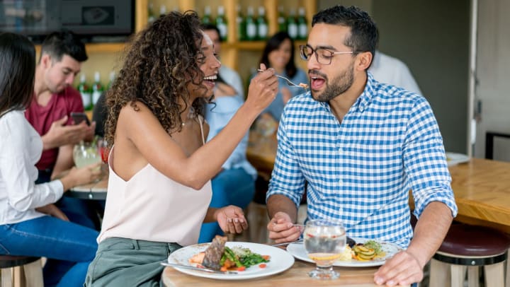 Woman feeding a bite of her food to a man with plates of food in front of them at a restaurant.