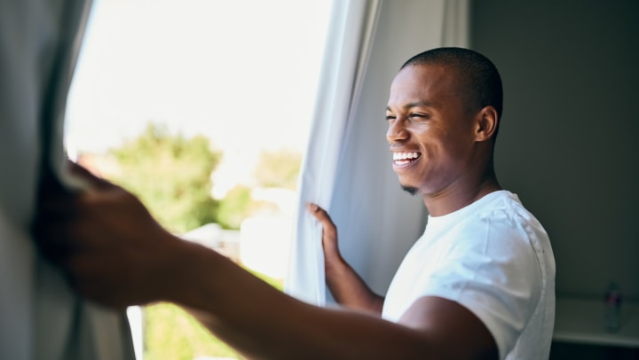 A young man opening his bedroom curtains in the morning.