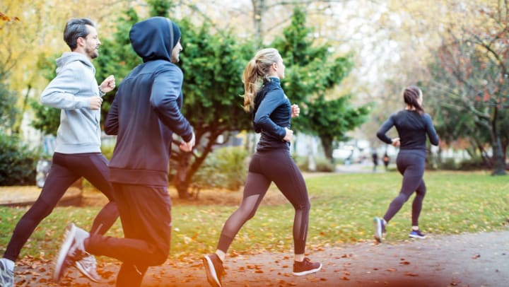A group of two young women and two young men wearing athletic clothes and jogging through a park in late fall