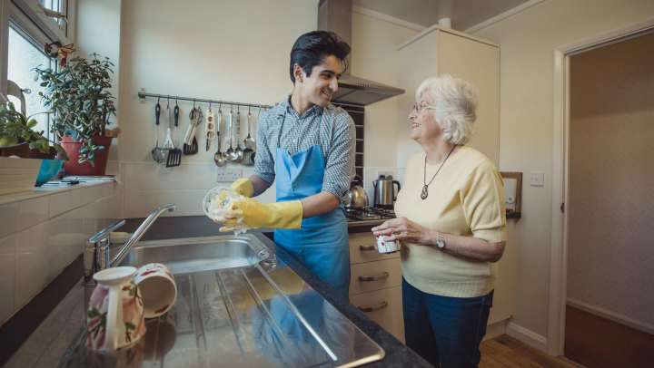 Two people standing in a kitchen, with one person washing dishes at the sink while wearing rubber gloves and an apron, and the other person holding a mug and standing nearby, with hanging utensils and countertops visible in the background
