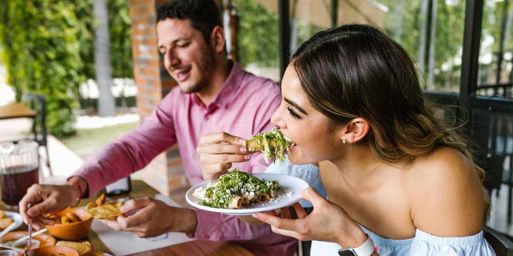 Couple enjoying a meal outdoors, woman taking a bite, man smiling with food on his plate at The Mezzanine at Freedom in Charlotte, North Carolina