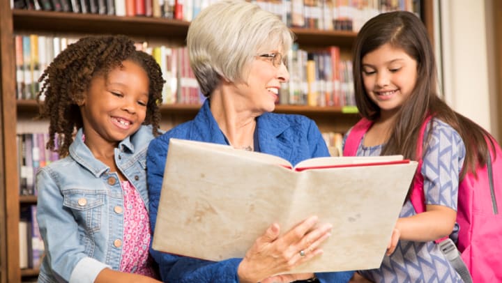Older woman in library looking at a textbook with two school-aged girls