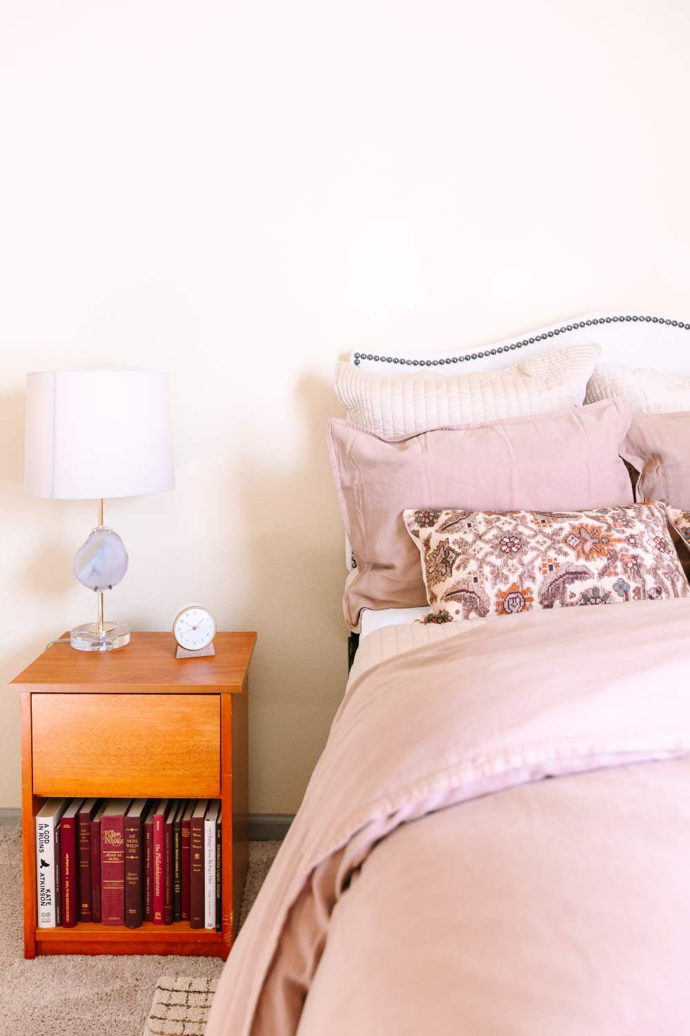 Bedroom bedside with wood nightstand, lamp, books, and layered pillows on bed at The Holly in Lubbock, Texas