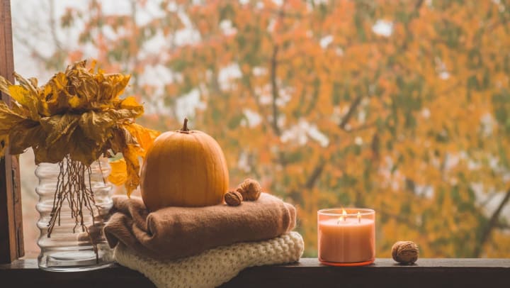 A window with blankets, a pumpkin, and a candle with an autumn tree in the background 