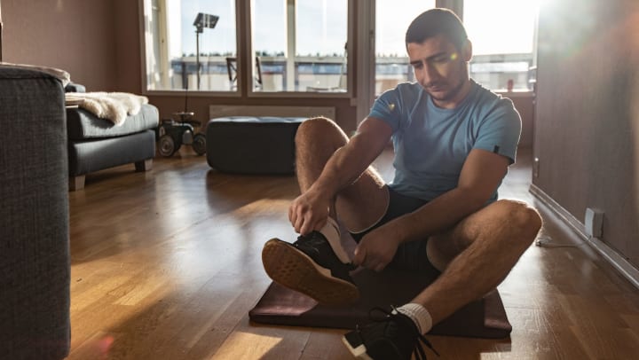 Young man putting on running shoes while seated on a yoga mat in a sunlit interior