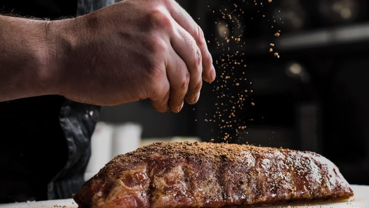 A hand sprinkling herbs and spices onto a piece of meat on a cutting board. 