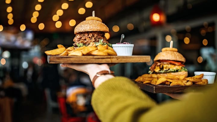 A server carries two wooden serving platters topped with burgers and pub fries through a busy Midland restaurant.