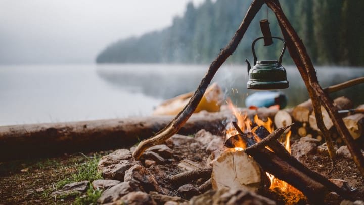 A green kettle hangs above a campfire in the foreground against a background of a calm lake and tall trees