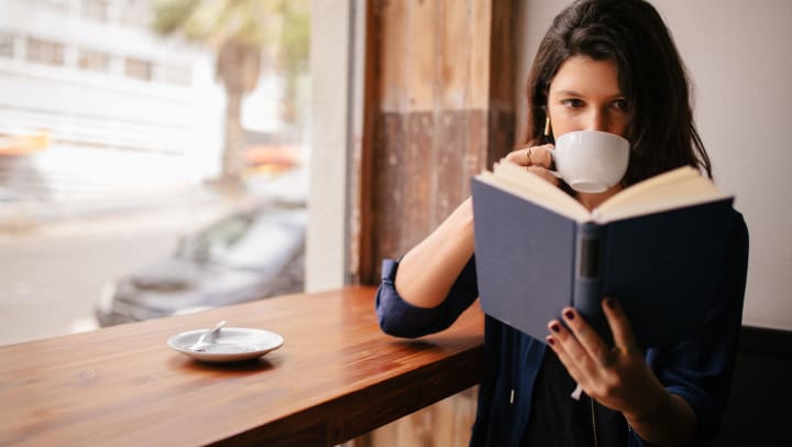 Woman sitting at a high bar with a white mug up to her mouth while she looks at an open book.