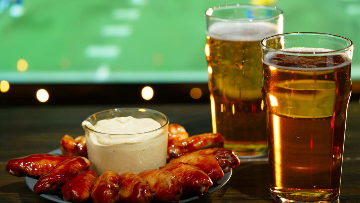 Plate of chicken wings and two beers in front of a sports television 