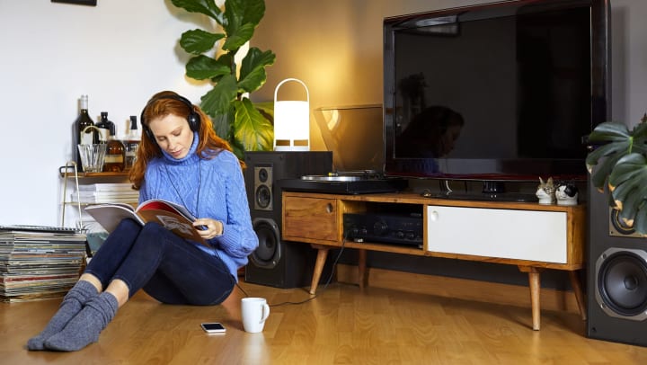 A woman sitting on the floor, listening to music and reading a magazine next to a TV that isn’t on. 