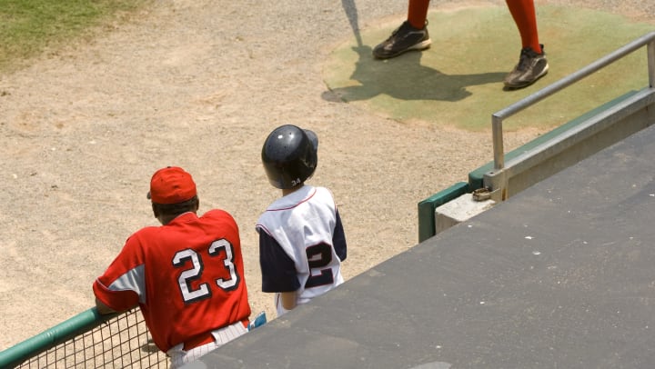 Baseball coach and bat boy standing in dugout at a game. A player is warming up in the on-deck circle nearby.