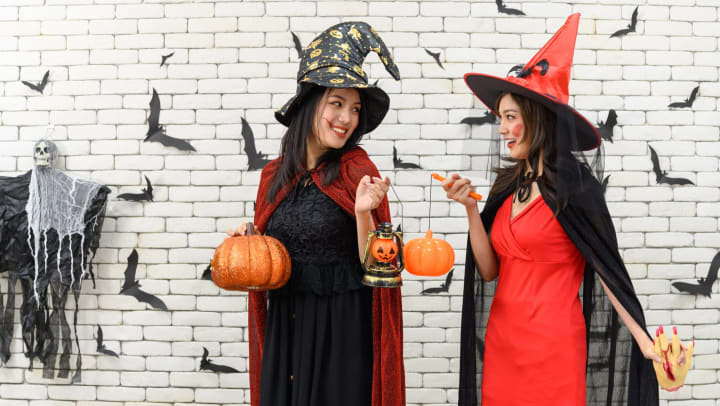 Two young women wearing black and red witch costumes and standing in front of a white brick wall with Halloween decorations 