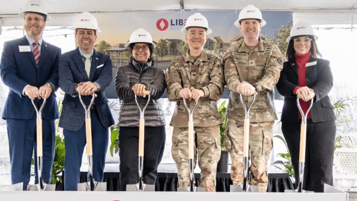 Liberty Military Team members and Joint Base Lewis McChord representatives stand in front of a Liberty Military Housing photo backdrop smiling with shovels in there hand and hardhats on to celebrate the Joint Base Lewis McChord Groundbreaking. 