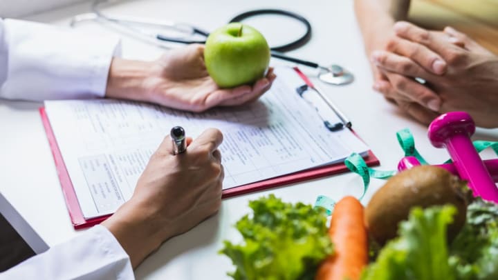 Close up of hands of a nutritionist and a patient during a consultation. The nutritionist is holding an apple while filling out paperwork and there are weights and vegetables on the table | nutritionists in savannah