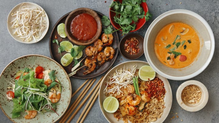 An overhead image of 8 plates of Thai food in varying sizes, including 4 main dishes of a soup, salad, shrimp, and pad Thai | Thai Restaurants Near Keller