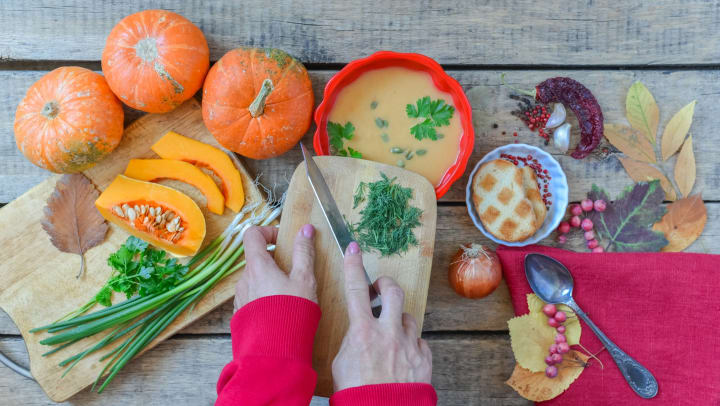 Hands holding a knife and a cutting board with herbs on it, surrounded by pumpkins, squash, and other seasonings.