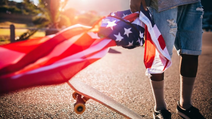 The bottom half of a man in rolled-up jeans and a gray sweatshirt. He’s holding an American flag that’s blowing in the wind and has a skateboard under his foot. 