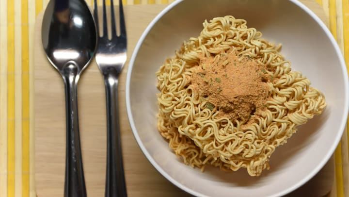 Bowl of instant noodles with the seasoning on top next to a fork and spoon.