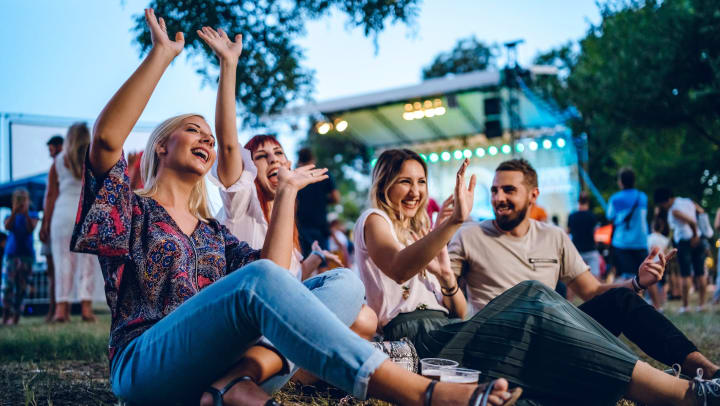 A group of four friends sitting on the grass enjoy live music at a festival. 
