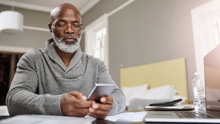 An older man looks at his mobile phone as he works on his finances at home, with a laptop and a calculator sitting nearby.
