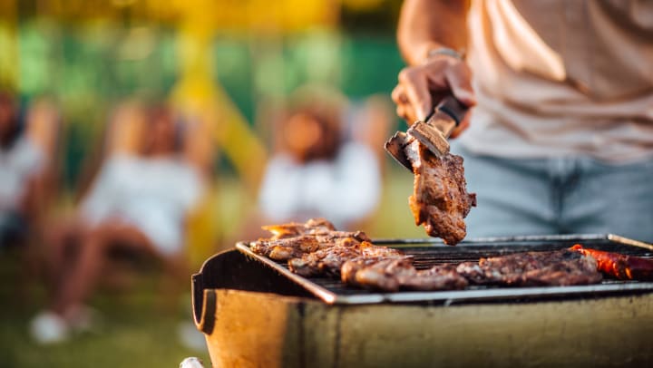 Hand holding tongs flipping a piece of meat on a barbecue. Background is blurred.