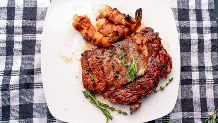 Ribeye steak with jumbo shrimp on a white plate sitting on a plaid linen cloth.