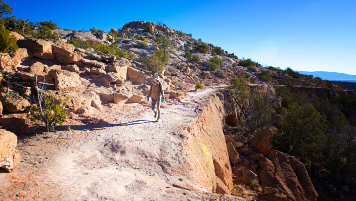 A hiker walking on a dirt trail near a cliff face under clear blue skies