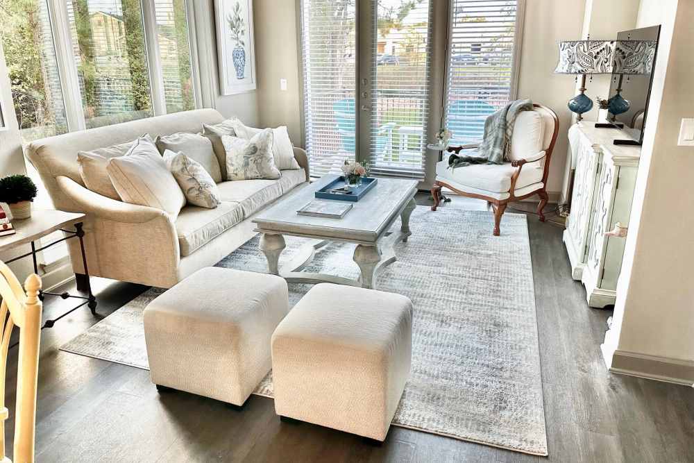 Cozy living room at Reserve Woodland Lakes with beige sofa, armchair, ottomans, large windows, and neutral tones.