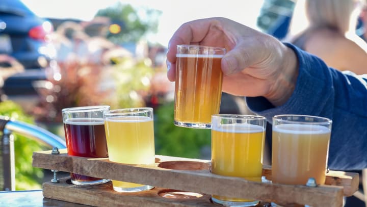Man outside, lifting a glass of beer out of a tasting tray full of different beers.