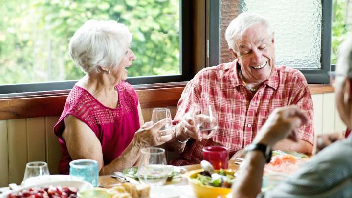 Two people seated at a dining table clinking glasses, with plates of food and drinks arranged in a bright indoor setting near large windows at {{location_name}} in {{location_city}}, {{location_state_name}}.