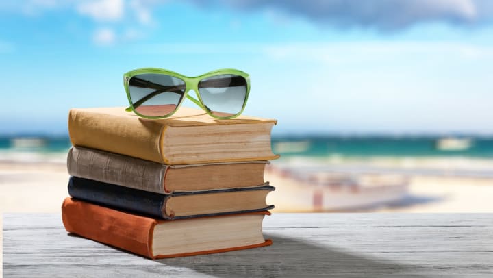Stack of books with sunglasses on top sitting on a wood table with the beach and ocean blurred in the background.