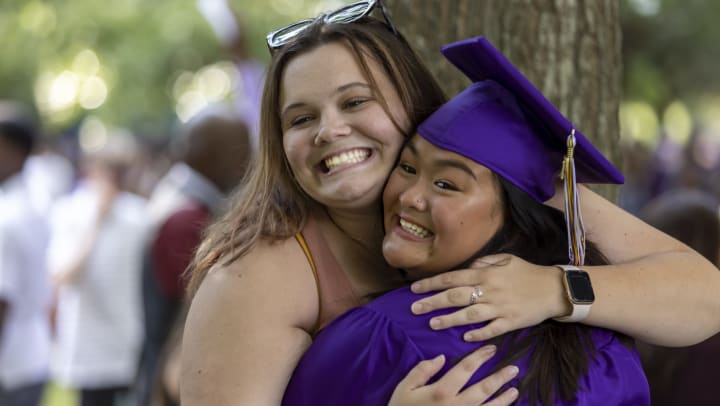 A smiling woman in a purple cap and gown hugs another woman. 