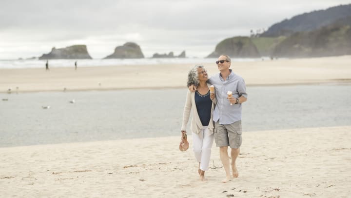 Elderly couple walking down the beach with ice cream cones.