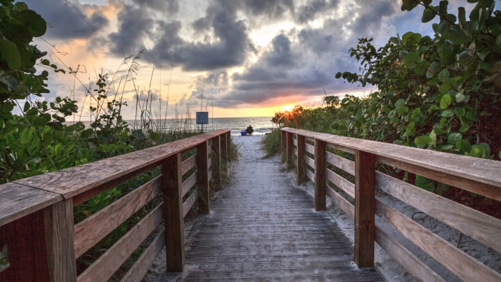 Photo taken from a wooden walkway leading out to the beach. In the foreground, there is a person sitting in a camping chair with a cooler at their side, looking out at the sunset.