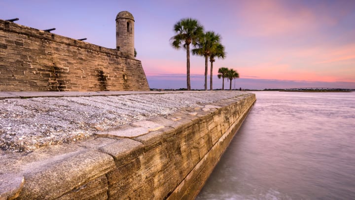 A view of the ocean at sunrise with a stone fort structure and palm trees