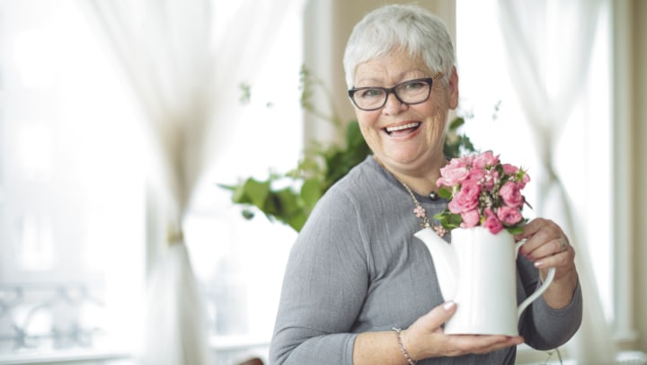 Senior woman holding a white pitcher with pink flowers in it and smiling.