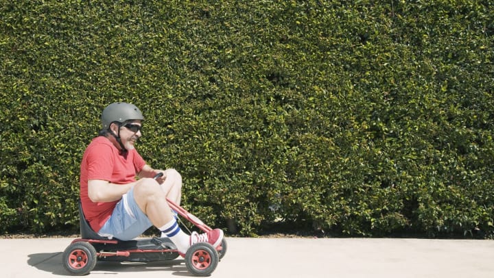 Man with a helmet on riding a tiny red go-kart on a sidewalk in front of  tall green hedges.
