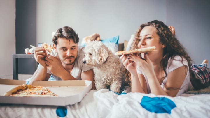 Young man and woman laying on a bed eating pizza, with an open pizza box sitting on the bed and a dog sitting between them