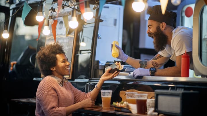 A man in a food truck putting mustard on a hot dog for a female customer. 