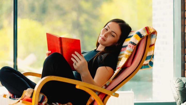 A young woman reads a book, sitting in a comfortable rocking chair with a colorful, striped blanket draped over it.