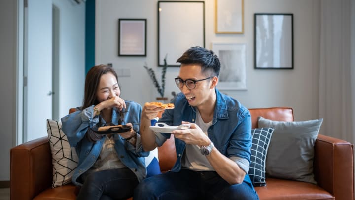 A young man and a young woman smiling, laughing, and eating pizza while sitting on a brown leather couch in an apartment with clean white walls