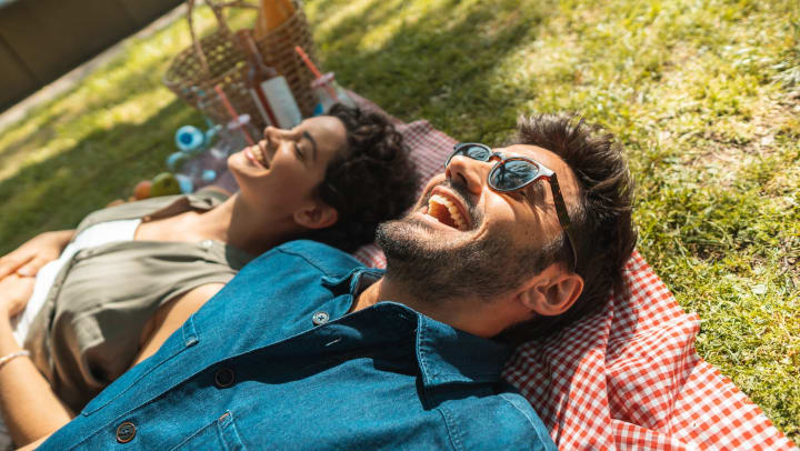Man and woman lying on a checkered blanket in the grass with smiles on their faces and a basket in the background.