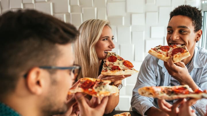 Three young people looking at one another, smiling and eating slices of pepperoni pizza. 