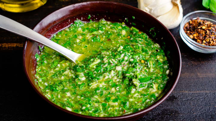 Bowl of chimichurri sauce with a spoon in it with a head of garlic, a pinch bowl of red chili flakes, a bunch of herbs, and a container of olive oil in the background.