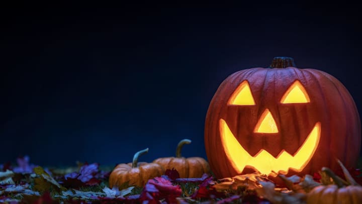 A smiling Jack O’ Lantern sitting in the grass with small pumpkins and fallen leaves at night for Halloween