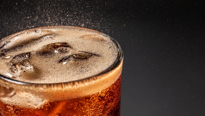 A glass of carbonated brown soda with ice against a dark grey background
