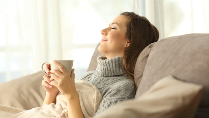 A woman with her eyes closed leaning back onto a sofa with a blanket and a coffee mug.